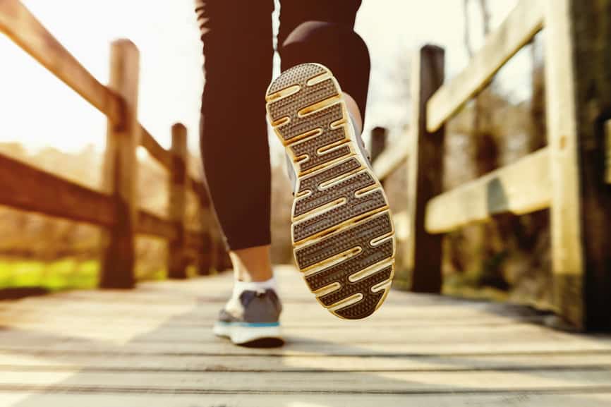 Close-up of a person running on a wooden bridge, focusing on the sole of their shoe. The background is slightly blurred, suggesting motion. The scene is outdoors with warm sunlight casting on the bridge and creating a dynamic, active atmosphere.