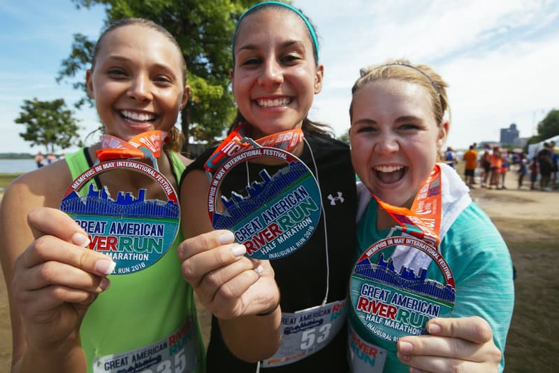 Three smiling women proudly display their medals after completing the Great American River Run marathon. They are outdoors, and other participants and trees are visible in the background. Each medal has a colorful ribbon and the event name prominently displayed.