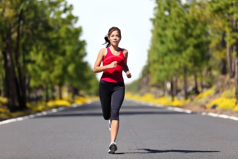 A person wearing a red tank top and black athletic pants is running on an empty road surrounded by green trees and yellow flowers. The scene is bright and clear, suggesting daytime in a natural, outdoor setting.