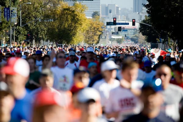 A large crowd of marathon runners fills a street under a sunny sky. Trees line the street, and a traffic light is visible in the background. The participants wear various athletic gear, hats, and sunglasses, with buildings in the distant background.