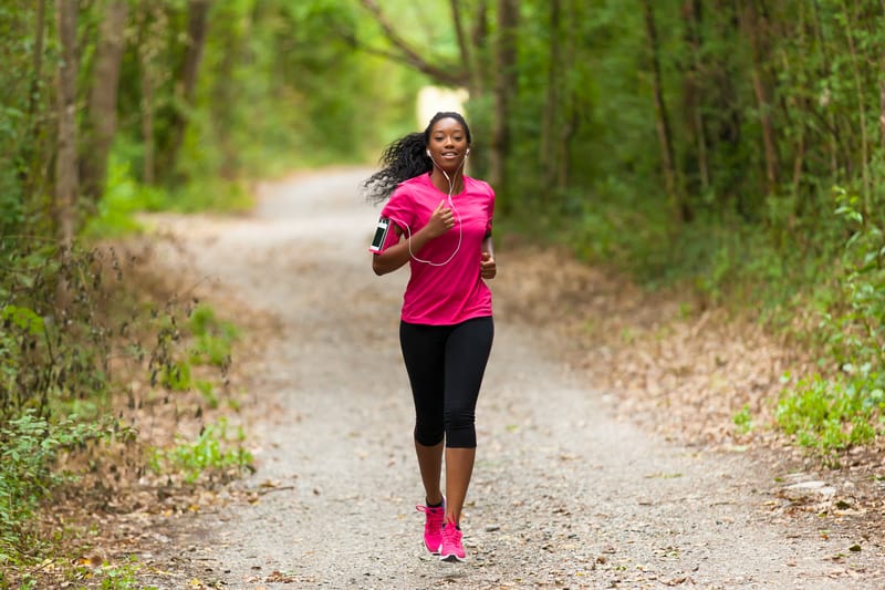 A person wearing a pink shirt and black leggings is jogging on a gravel trail through a lush, wooded area. They have a smartphone strapped to their arm and are smiling as they run. The path is surrounded by green trees and vegetation.