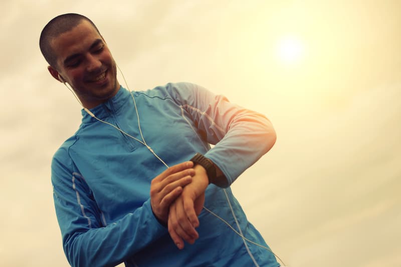 A man in a blue long-sleeve athletic top stands outdoors under a bright sky, smiling as he looks at a watch on his wrist. He has earphones in his ears, and the sun is setting in the background.