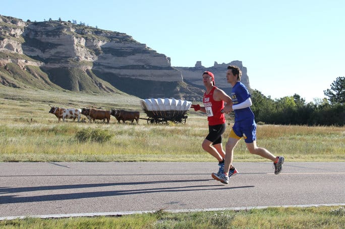 Two runners in red and blue outfits run on a road by a scenic landscape featuring rocky formations, grassy fields, and an old-fashioned wooden wagon. Cows graze nearby under a clear, sunny sky, creating an atmosphere of a countryside race.