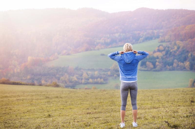 A person in athletic wear stands on a grassy hill, facing away, looking at a vast landscape of rolling hills and trees under a slightly hazy sky. They are adjusting the hood of their blue sweatshirt. It appears to be early morning or late afternoon.