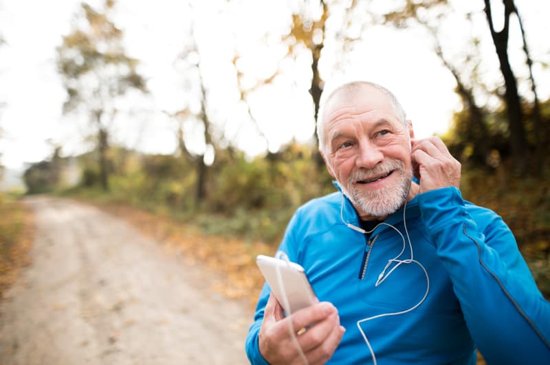 An older man with a beard wearing a blue pullover is smiling and wearing earphones while holding a smartphone. He is standing on a dirt path lined with trees. The background has autumn foliage, giving the scene a peaceful, outdoor ambiance.