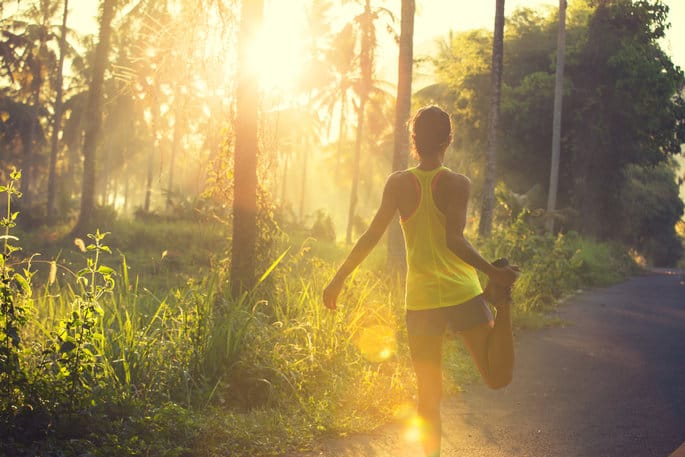 A person in athletic wear, illuminated by the warm sunlight, stretches one leg while standing on a paved path surrounded by lush greenery and tall trees. The sun is low in the sky, casting a golden glow over the scene.