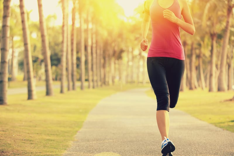 A person in a pink tank top and black leggings jogging on a paved path lined with palm trees. The sun is shining brightly, casting a warm glow over the scene.