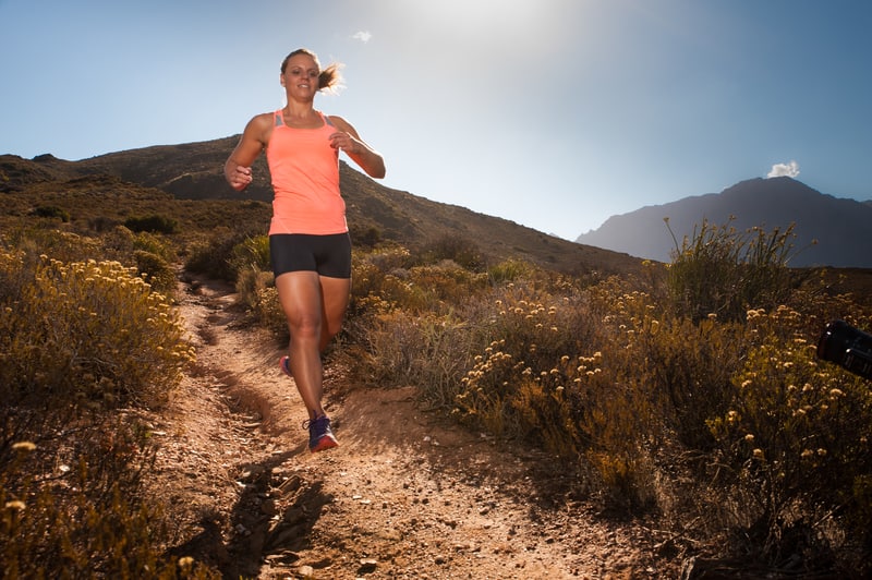 A woman in an orange tank top and black shorts jogs down a narrow dirt trail through a scenic, sunlit landscape with dry shrubs and mountains in the background. The sky is clear and bright, suggesting a sunny day.