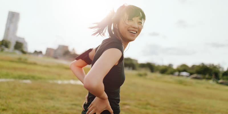 A woman with a ponytail is jogging in a grassy field on a sunny day. She is wearing a black shirt and smiling at the camera. Trees and a few buildings are visible in the background.