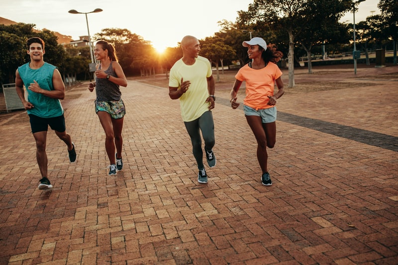 Four people run outdoors on a brick pathway at sunrise or sunset. They are dressed in athletic wear, with one person wearing a cap. Trees and park elements are visible in the background, and everyone appears to be enjoying the run while chatting.