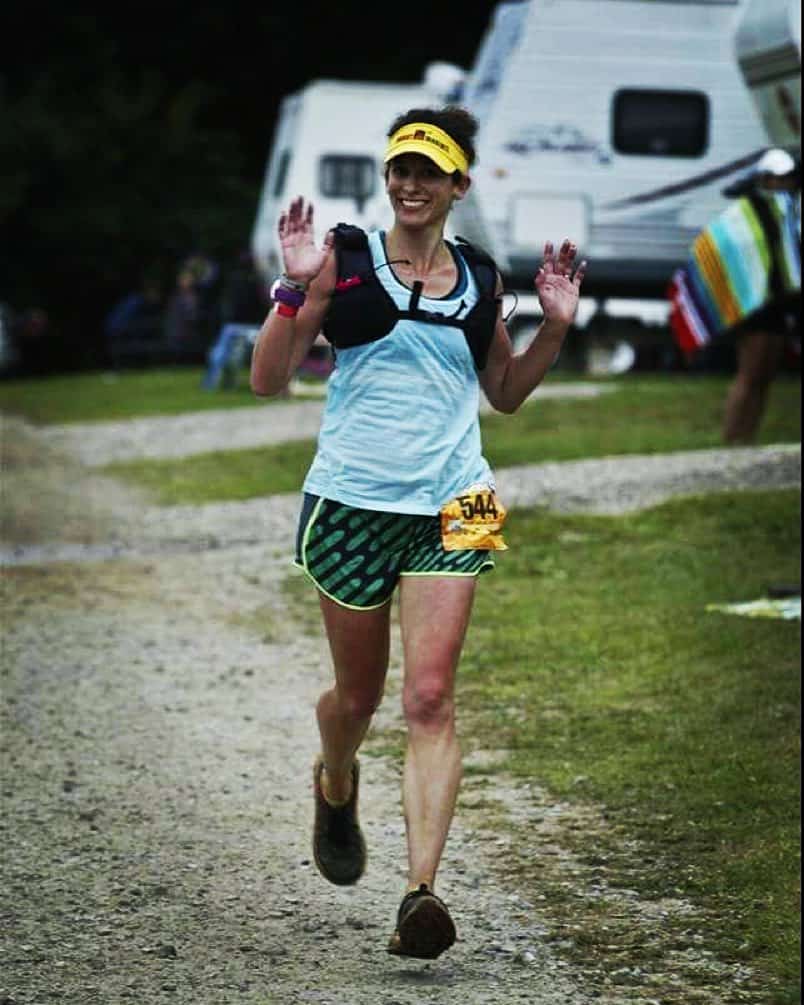 A person wearing a blue tank top, patterned shorts, and a yellow visor smiles and waves while running on a gravel path. They have a race bib with the number 541 attached to their shorts and are in front of a white RV and a colorful striped towel in the background.