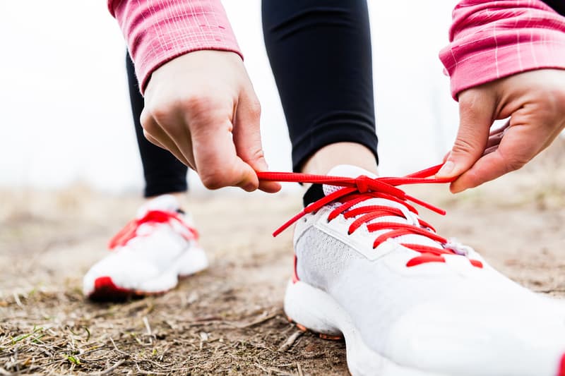 A person in black leggings and a pink long-sleeve top ties the bright red laces of their white running shoes. The scene is outdoors on a dirt surface.