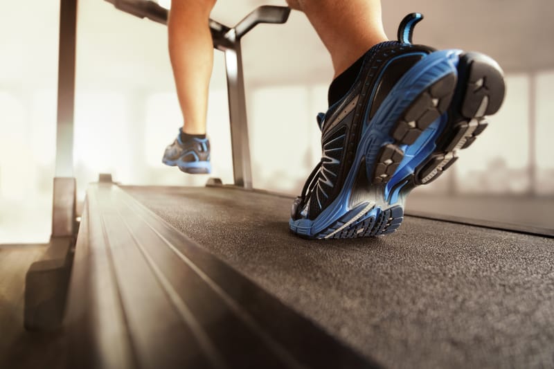 Close-up shot of a person’s legs and feet running on a treadmill. The individual is wearing black and blue athletic shoes. The background is blurred, creating a sense of motion and focus on the running action.