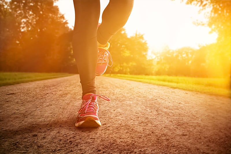 A close-up of a person running on a dirt path surrounded by greenery, taken during sunset or sunrise. The runner is wearing bright pink running shoes and black leggings, and the scene is bathed in a warm, golden light.