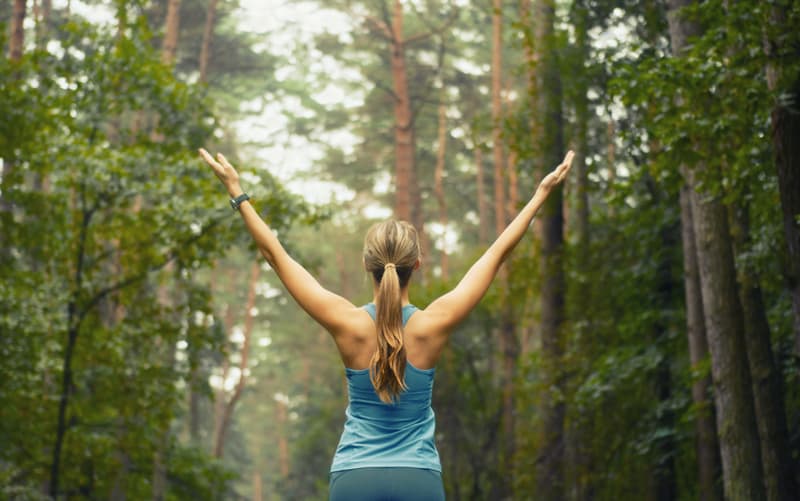 A person with long hair in a ponytail, wearing a blue tank top, stands with arms raised in the middle of a dense, green forest. The scene conveys a sense of freedom and connection with nature.