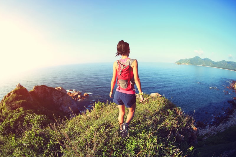 A person with a ponytail, wearing a red tank top, blue shorts, and a red backpack, is standing on a grassy cliff overlooking the ocean. Rocky outcrops and a distant mountainous coastline are visible under a clear blue sky.