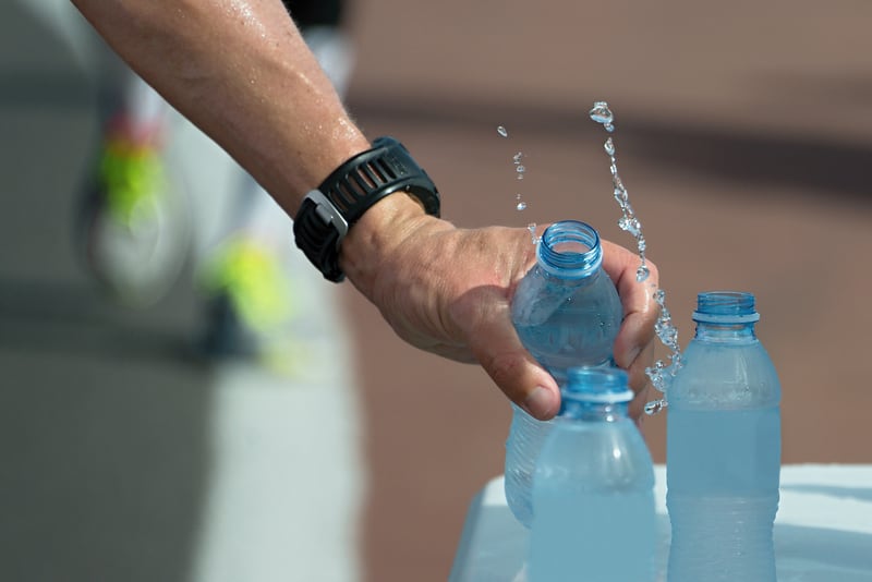 A close-up of a person's hand, wearing a black wristwatch, pouring water from one blue plastic bottle into another. A splash of water is visibly mid-air between the bottles. Two other blue plastic water bottles are placed on a surface nearby.