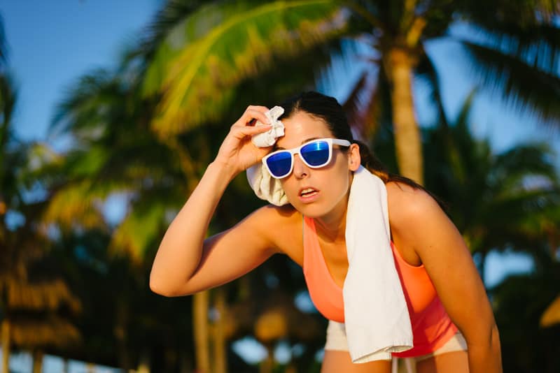 A woman wearing sunglasses and a peach tank top, with a white towel draped over her shoulder, wipes sweat from her forehead with a small cloth. She is outdoors, with palm trees and a clear blue sky in the background, suggesting a hot, sunny environment.