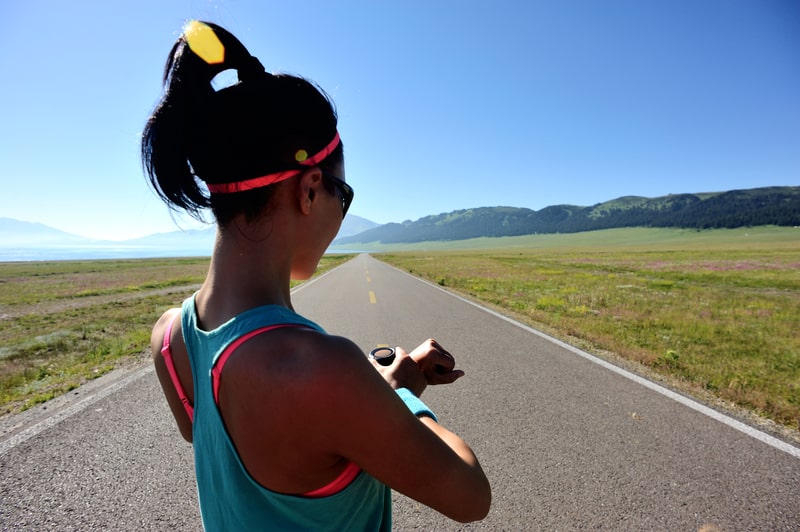 A person wearing a tank top and sunglasses checks their smartwatch while standing on an open, deserted road surrounded by green fields and distant mountains under a clear blue sky. The person's hair is tied up in a ponytail, and they appear to be in a running stance.