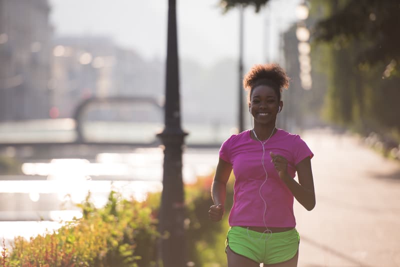 A person wearing a pink shirt and green shorts jogs along a sunny, tree-lined path by a body of water. They are smiling and have earbuds in, enjoying the morning exercise. The background features a blurred bridge and buildings.