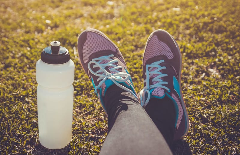 A person wearing pink and gray running shoes with blue laces is sitting on grass. A white water bottle is placed on the grass to the left of the shoes. The scene is bathed in warm sunlight.