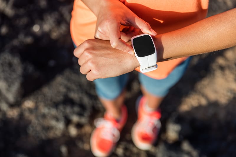 A person wearing a bright orange shirt and blue shorts adjusts a white smartwatch on their wrist. The background is out of focus, and the person is also wearing bright orange sneakers. The scene appears to be outdoors.