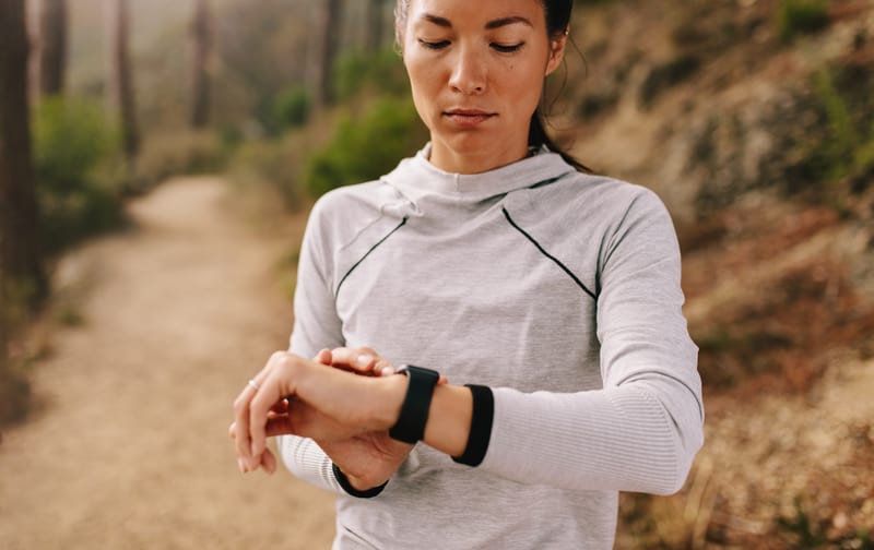 A woman on an outdoor trail looks down at her smartwatch while wearing a light-colored long-sleeve athletic top. She appears focused, with trees and foliage visible in the background. The path she's on suggests a nature or forest area ideal for running or walking.