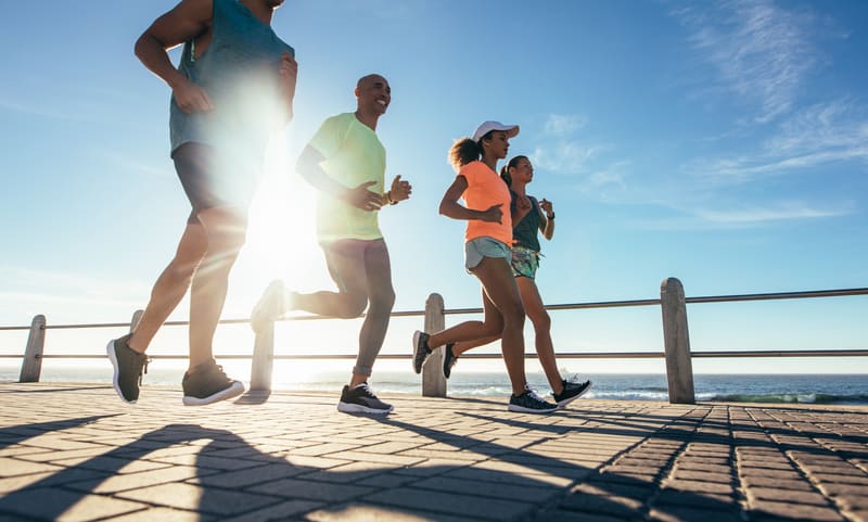 Four people jog along a seaside promenade with a bright blue sky and the sun shining. They are dressed in athletic wear, with the ocean visible in the background and railings lining the path. The scene is energetic and vibrant, highlighting a group run by the coast.