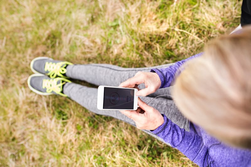 A person wearing a purple long-sleeve shirt, gray leggings, and gray sneakers with yellow laces sits on a grassy field while looking at a smartphone. The person appears to be using an app, and only their upper torso, legs, and feet are visible in the frame.