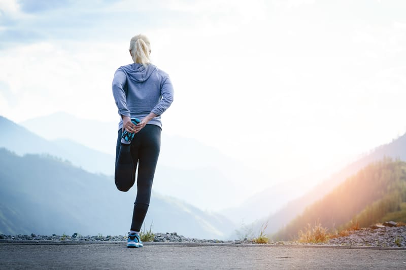 A person with blonde hair, wearing a hoodie and leggings, stretches their leg while standing on a mountain road. The person faces away, with a scenic view of mountains under a bright, hazy sky in the background.