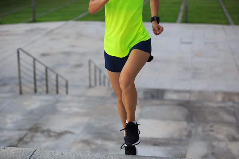 A person in a bright yellow shirt and dark shorts is jogging up concrete steps. The individual is wearing a wristwatch and black athletic shoes. Green grass and railing are visible in the background.