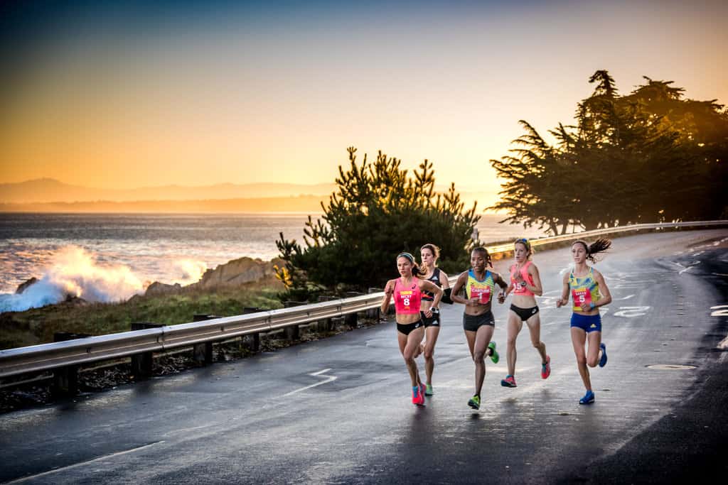 A group of five runners in vibrant athletic gear race along a coastal road at sunrise, with waves crashing against the shore and trees silhouetted in the background. The road and ocean create a picturesque, dynamic setting for the competition.