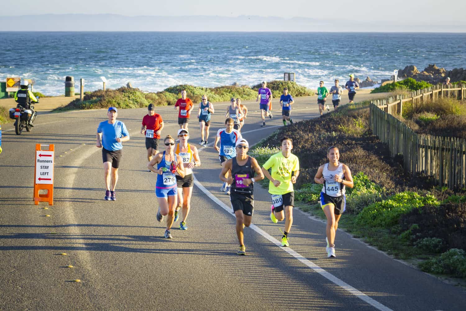 A group of runners participates in a road race along a coastal highway with the ocean in the background. They are wearing various race bibs and athletic gear. A traffic cone and a sign indicating course directions are visible on the road. The weather looks clear and sunny.