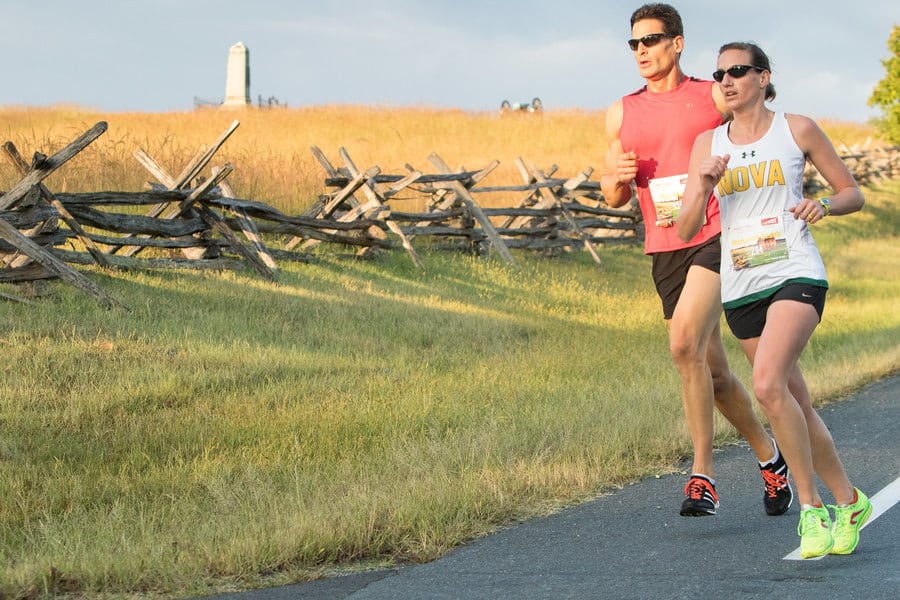 Two runners are on a paved path next to a split-rail fence in a rural area with tall grass. One wears a red sleeveless top and black shorts, the other a white tank top with "NOVA" printed on it, and black shorts. Both are running in the same direction.