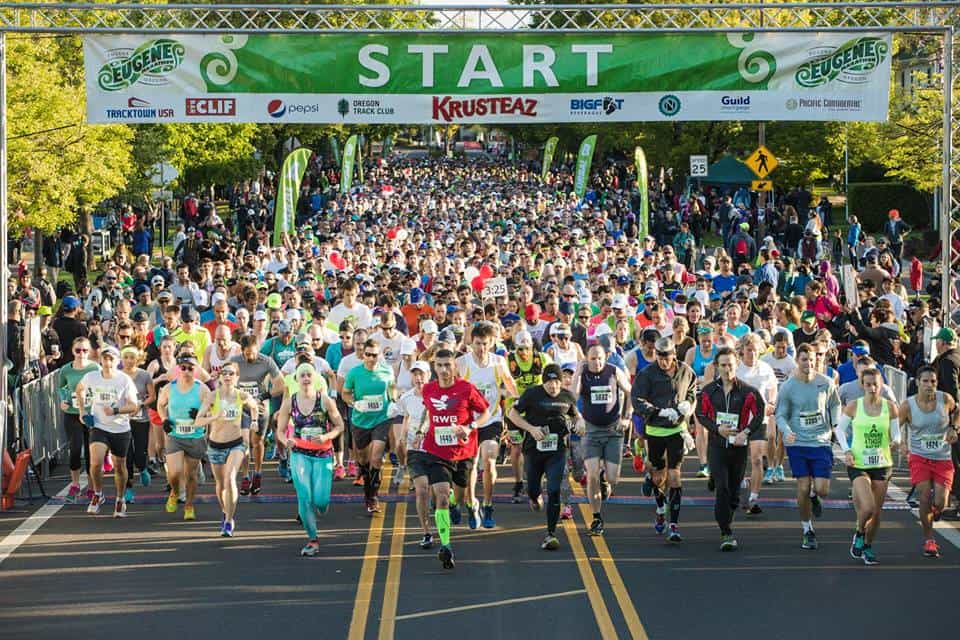 A large crowd of runners begins a race, passing under a "START" banner adorned with various sponsor logos. Participants are dressed in colorful athletic wear, and the weather appears sunny with green trees in the background.