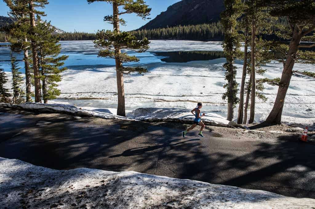 A lone runner jogs along a paved road through a forested area beside a partially frozen lake. Tall trees surround the scene, with mountains in the background beneath a clear blue sky. Snow patches and the lake's ice create a wintry contrast to the runner’s movement.