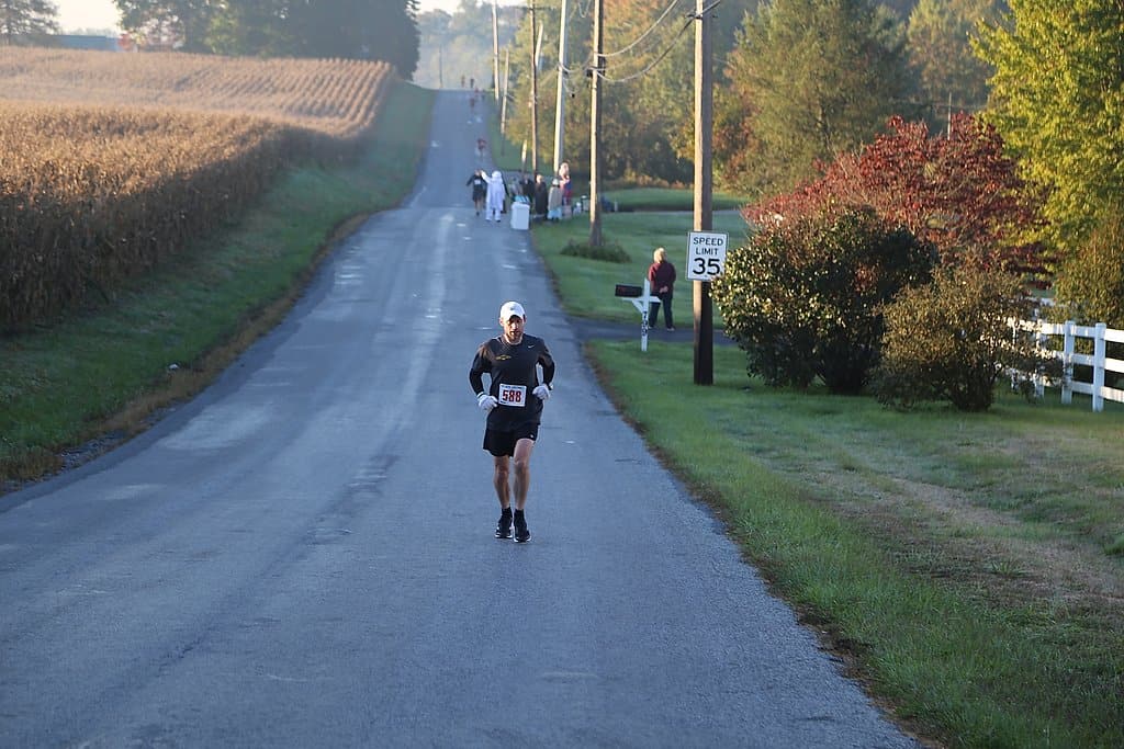 A runner wearing a black outfit and a white cap jogs down a country road during a race. Surrounding him are fields and trees, some with autumn colors. There are a few spectators and other runners in the distance. A speed limit sign reads 35 mph.