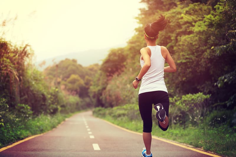 A person wearing a white tank top and black leggings is jogging on a paved road surrounded by lush greenery, with trees lining both sides. The sun is setting, casting a warm glow over the scene. The person’s hair is in a ponytail, and they are wearing a headband.