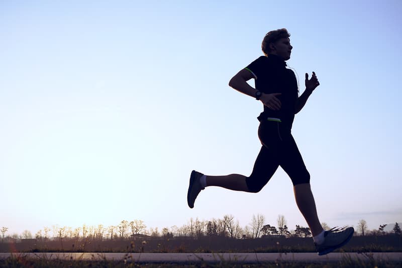 Silhouette of a person running outdoors on a flat path during sunset or sunrise, with trees and a clear sky in the background. The person is wearing athletic clothing and appears to be engaged in a focused, vigorous run.