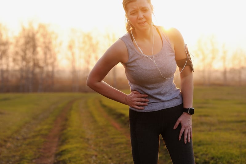 A woman wearing a gray tank top and black leggings, with earphones and a smartwatch, stands outdoors on a grassy path. She appears to be in discomfort, holding her side with one hand and leaning forward slightly, with a pained expression on her face.