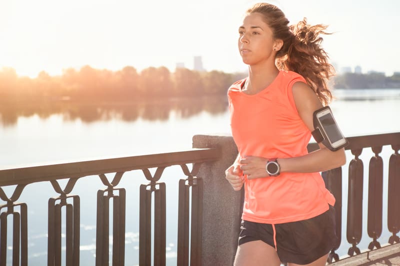 A woman jogs along a waterfront path during sunrise or sunset. She is wearing an orange sleeveless top, black shorts, and has a smartphone strapped to her arm. A watch is visible on her wrist. Tree line and city buildings can be seen in the background.