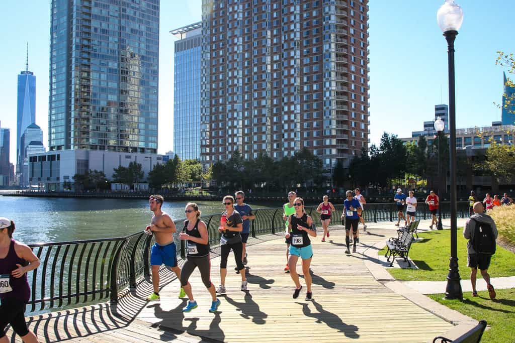 A group of runners participating in a race jog along a waterfront path lined with tall buildings and trees. The sun is shining, casting shadows on the path, and some spectators watch from the side. Participants wear race numbers pinned to their clothes.