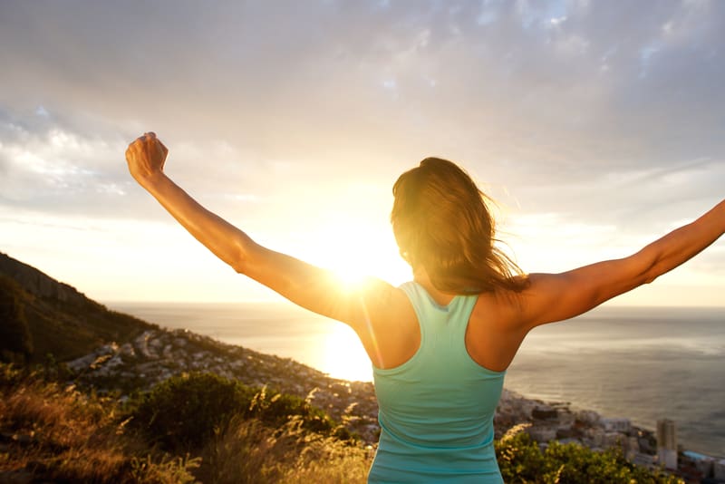 A person in a blue tank top stands with arms raised, back facing the camera, overlooking a scenic coastal landscape at sunset. The sun is partially hidden by clouds, casting a warm glow over the ocean and nearby terrain.