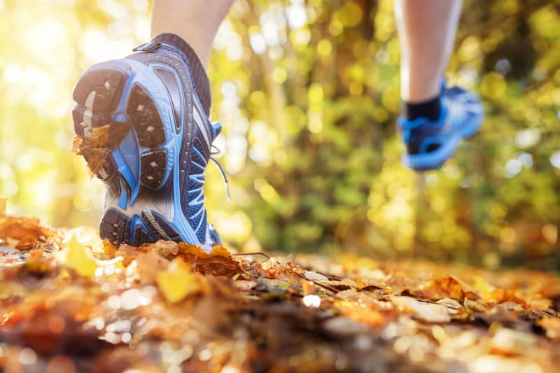 A person running in a forest with blue running shoes. The trail is covered in fallen autumn leaves, and sunlight filters through the trees, creating a warm and vibrant atmosphere. The image is focused on the runner's shoes and the ground.