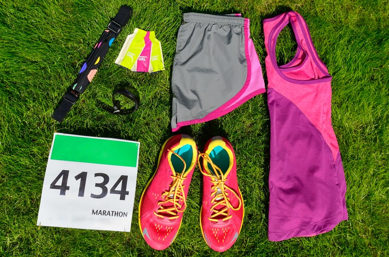 Top view of marathon race gear laid out on grass. Includes a race bib numbered 4134, pink and yellow running shoes, a pink and purple athletic tank top, gray and pink shorts, an armband, a sports watch, a colorful headband, and a packet of energy gel.