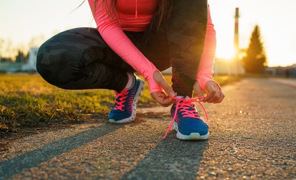 A person with long hair, wearing a bright pink long-sleeve shirt and black camouflage leggings, is crouched down on a paved path at sunset, tying the laces on their blue running shoes with pink laces.