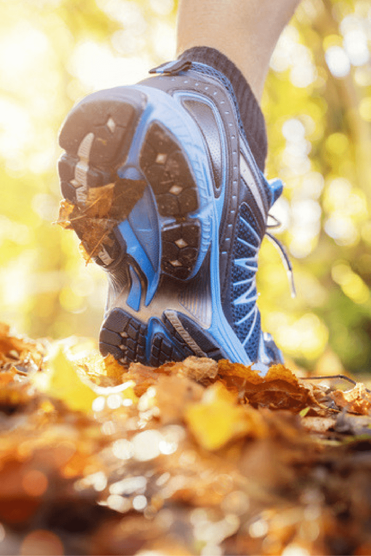 Close-up of a person wearing blue running shoes walking on a forest trail covered with autumn leaves. The sun shines through the trees, creating a warm, golden light. The shoes are stepping on the leaves, and one leaf is caught in the shoe tread.