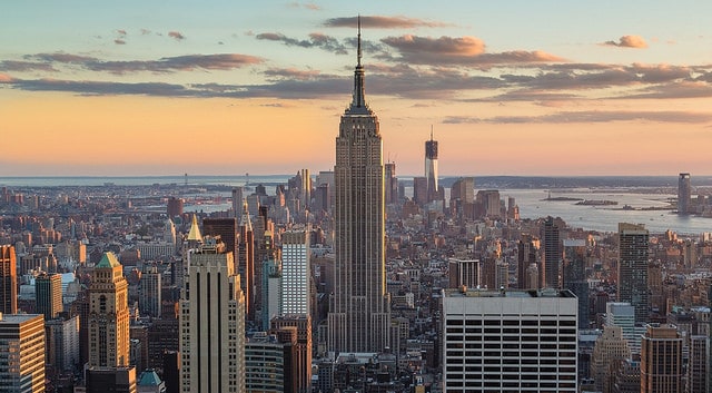 Aerial view of a city skyline at sunset, featuring a prominent skyscraper in the center. The buildings are illuminated by the warm glow of the setting sun, with a river and bridges visible in the background and scattered clouds in the sky.