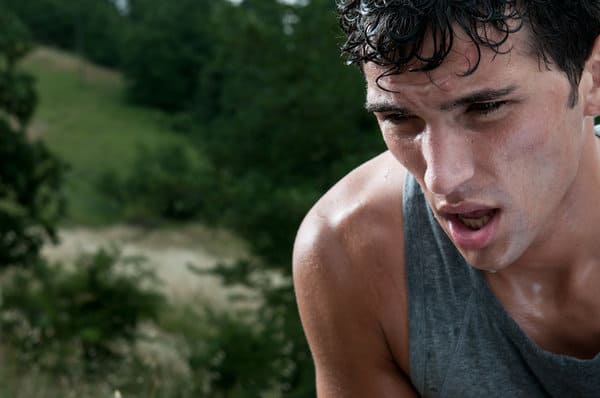 A young man with sweat on his face and wet hair appears exhausted as he catches his breath. He is wearing a sleeveless gray shirt and is outdoors, with a blurred background of greenery and trees.