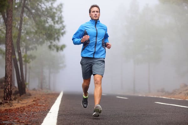 A person in a blue jacket and gray shorts is jogging on an empty, foggy road. The background is misty with trees lining the sides of the road. The scene evokes a sense of calm and focus.